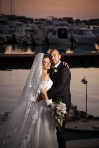 Portrait of smiling bride and groom embracing against river at dusk