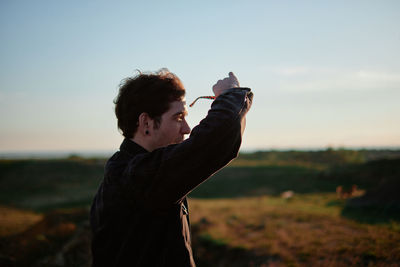 Side view of man holding sunglasses looking away while standing against landscape