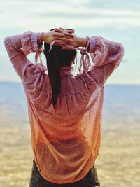 Rear view of woman with hands behind head standing at beach against sky