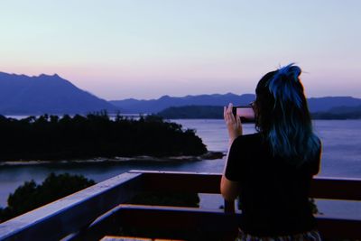 Woman standing on railing by lake against sky during sunset
