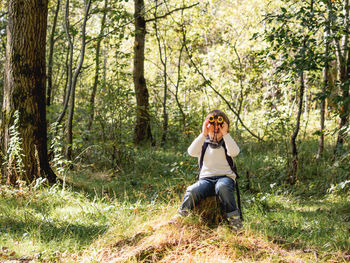 Little explorer on hike in forest. boy with binoculars sits on stump.outdoor activity for children. 