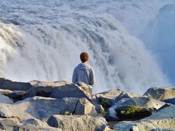 Rear view of man sitting on rock against waterfall