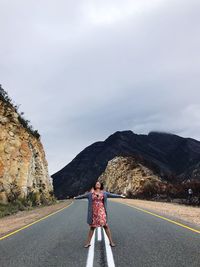 Mature woman with arms outstretched standing on road against cloudy sky