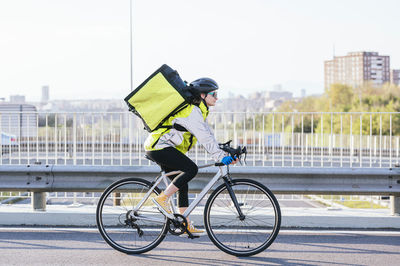 Side view of female courier with thermal bag riding bike on bridge while delivering food in city