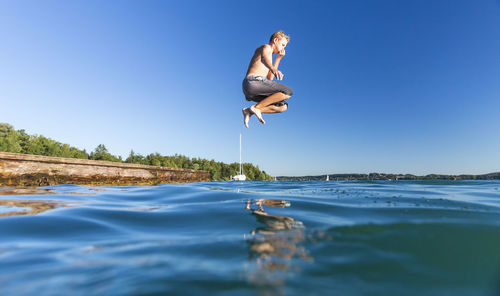 Full length of shirtless boy jumping over lake against clear blue sky