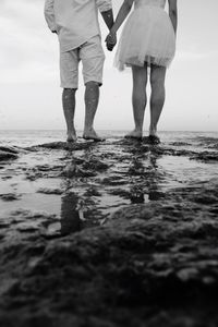 Low section of couple holding hands while standing on rock by sea