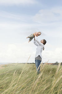 Side view of woman holding aloft baby on grassy field against sky