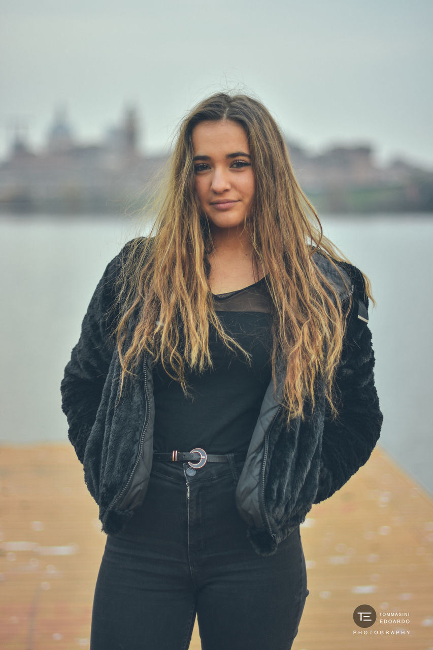 PORTRAIT OF SMILING YOUNG WOMAN STANDING AGAINST THE SEA