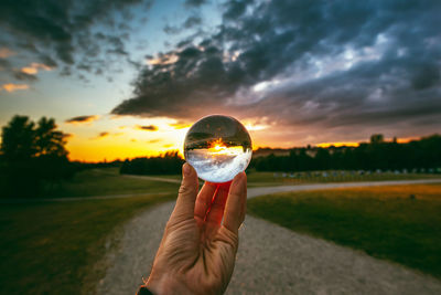 Cropped person holding crystal ball against cloudy sky during sunset