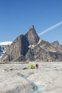 Two mountaineers stand next to tent on glacier under mountain summit.