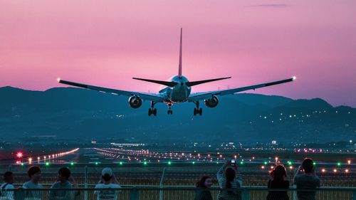 People by railing with airplane flying in sky at night