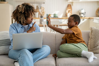Freelancer black mother working on laptop at home, noisy african child distracts asking attention.