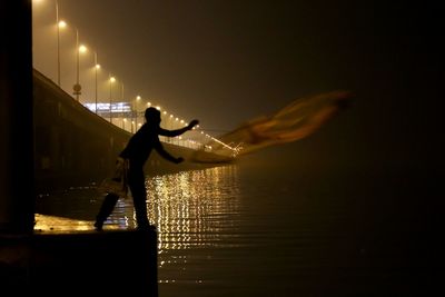 Silhouette fisherman throwing net into sea by illuminated penang bridge at night