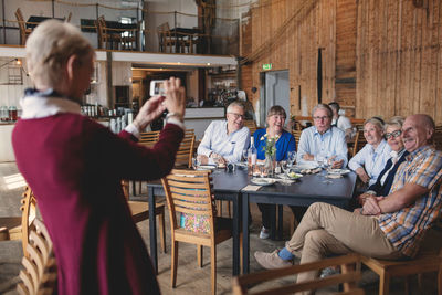 Senior woman photographing happy friends sitting at table in restaurant