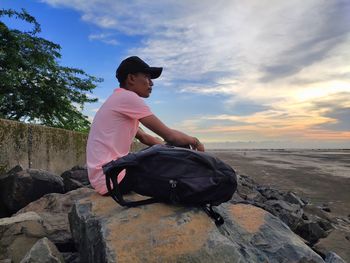 Man sitting on rock against sky during sunset