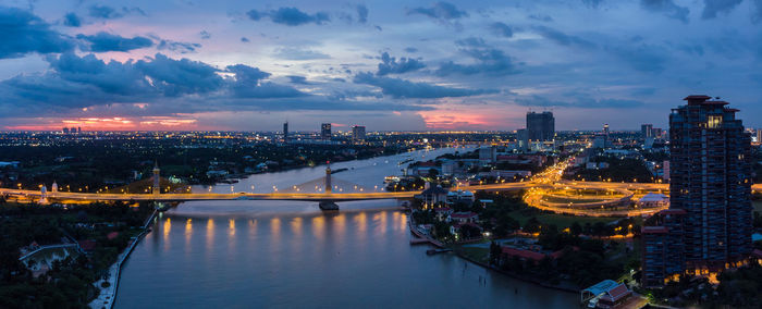 Illuminated bridge over river by buildings against sky at sunset