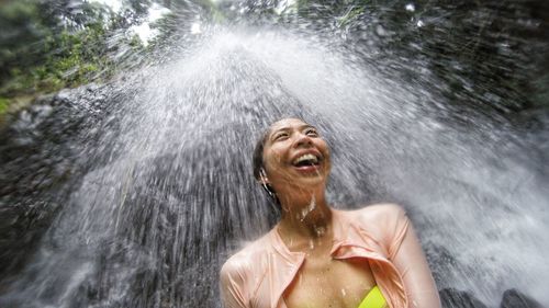 Low angle view of cheerful woman standing below waterfall