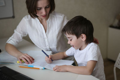 Beautiful mother teaches her son at home at the table, real interior