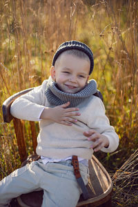 Boy in warm clothes sit on chair along a path on a field with dried grass in autumn