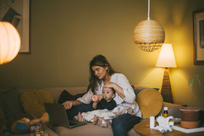 Baby girl with bottle sitting by mother in living room during pandemic
