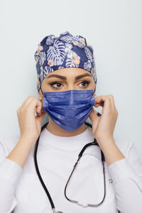 Doctor woman wearing medical white robe and uniform bandana with stethoscope on neck fixing protective mask on face looking away
