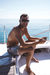 Portrait of shirtless man wearing sunglasses while sitting in boat on sea against clear sky during sunny day