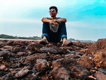 Young man sitting on rock by sea against sky