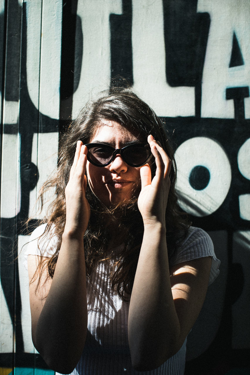 PORTRAIT OF YOUNG WOMAN WEARING SUNGLASSES AGAINST WALL