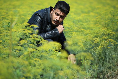 Man wearing black leather jacket crouching amidst plants on field during sunny day