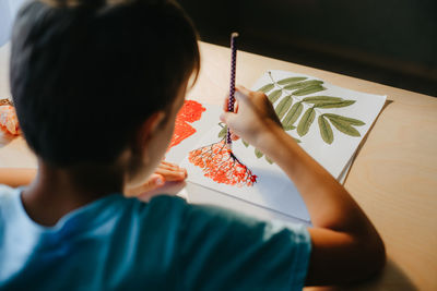 Cute child boy siting at desk and drawing rowanberries on album sheet with dry rowan leaves. 