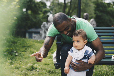 Father pointing and showing to toddler while sitting at bench in park