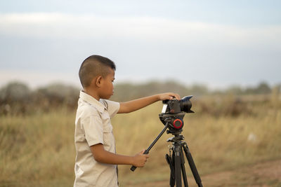 Side view of boy photographing while standing on field against sky