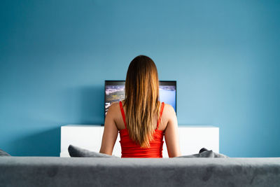 Rear view of woman looking at television set against blue wall at home