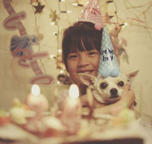 Portrait of cute smiling girl holding dog while sitting by birthday cake at home