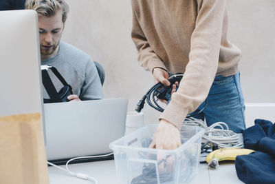 Midsection of female computer programmer removing cables from container while colleague holding headphones in office