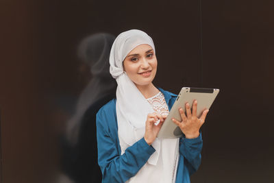 Portrait of young woman holding digital table against black background