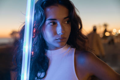 Thoughtful millennial long haired brunette looking away pensively while standing on illuminated street in summer evening