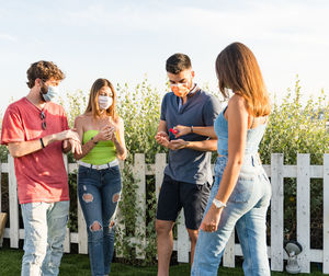 Young friends wearing mask using hand sanitizer while standing by railing