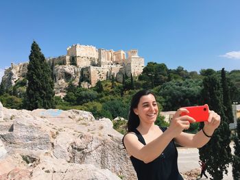 Happy woman taking selfie while standing by rocks against acropolis of athens