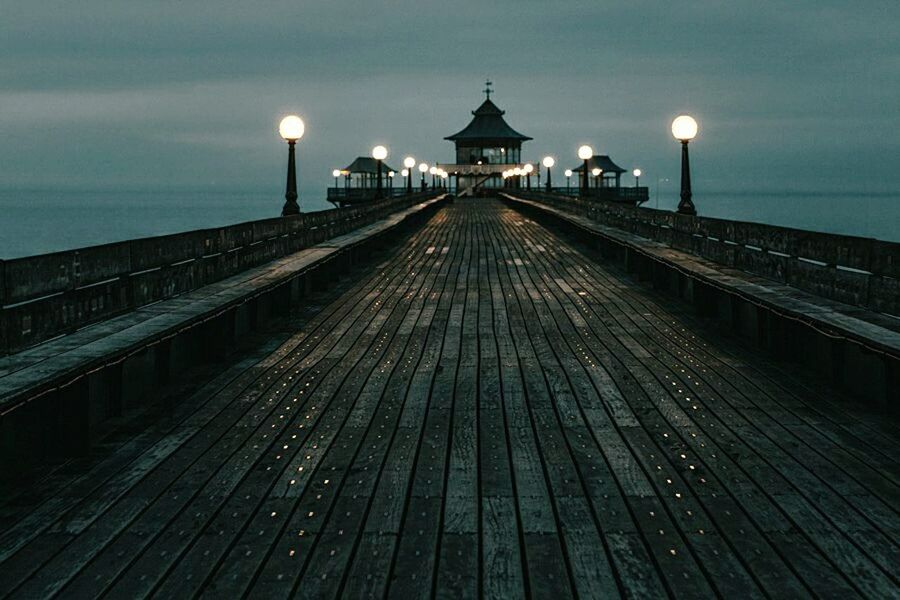 ILLUMINATED WALKWAY BY SEA AGAINST SKY AT DUSK