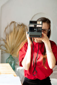 A young woman in a red t-shirt holds a polaroid in her hands and takes a photo