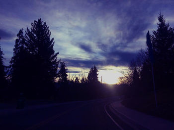 Road amidst silhouette trees against sky at sunset