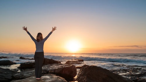 Rear view of woman exercising yoga with hands raised on rock at beach against sky during sunrise