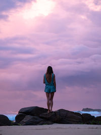 Rear view of woman standing on rock against sky during sunset