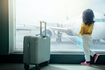 Full length rear view of girl standing by luggage while looking through window at airport