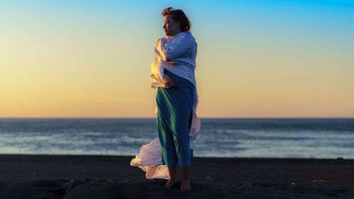 Full length of woman with fabric standing at beach during sunset against sky