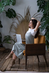 Beautiful woman with hands behind head and computer on knees resting, daydreaming in surround plants