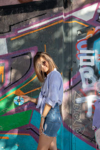 Beautiful woman holding compact disc while standing against graffiti wall