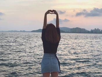 Rear view of woman making heart shape with fingers against sky