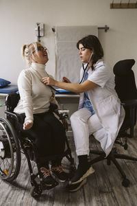 Full length of female healthcare worker examining disabled patient with stethoscope in medical clinic